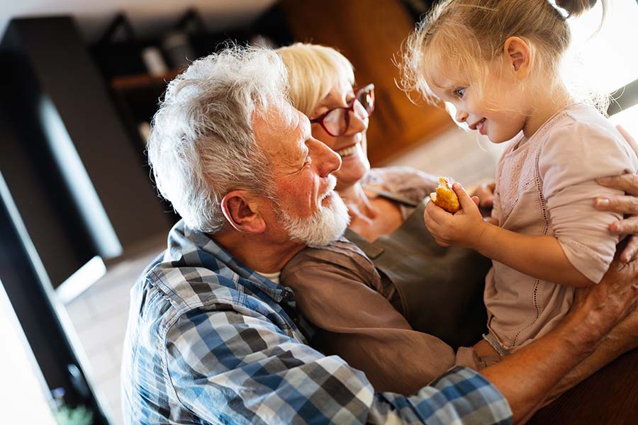 About Our Agency - Portrait of Cheerful Grandparents Playing with Their Granddaughter in the Kitchen as She Eats a Piece of Bread
